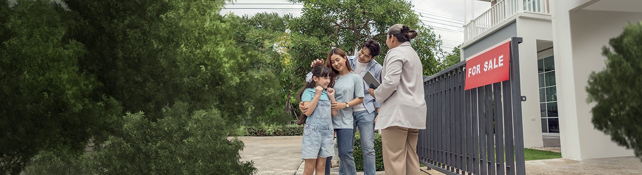 Female house salesman, home broker, standing in a suit, standing in front of a house for sale sign. There are family, parents, daughters coming to visit the project asking for details of the house for