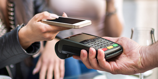 One of two female customers is holding mobile phone over credit card reader for contactless payment transaction in the coffee shop. Coffee cups and glasses are on the table.